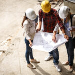 Two female inspectors and architects discuss with head engineer about blueprints of construction site.